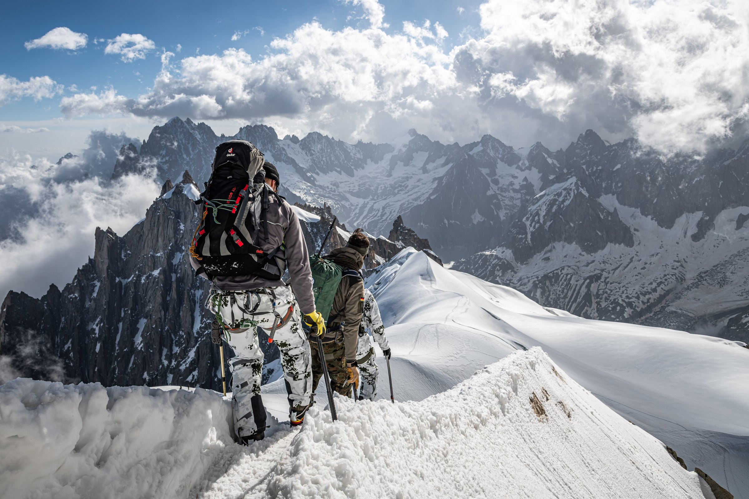 Drei Bergsteiger in Tarnkleidung gehen hintereinander über einen schmalen Schneegrat, dahinter Gipfel und Wolken