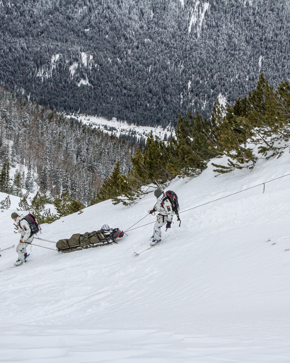Soldaten in Schneetarn auf Ski ziehen einen Transportschlitten mit einem Verwundeten an Seilen über einen steilen Hang