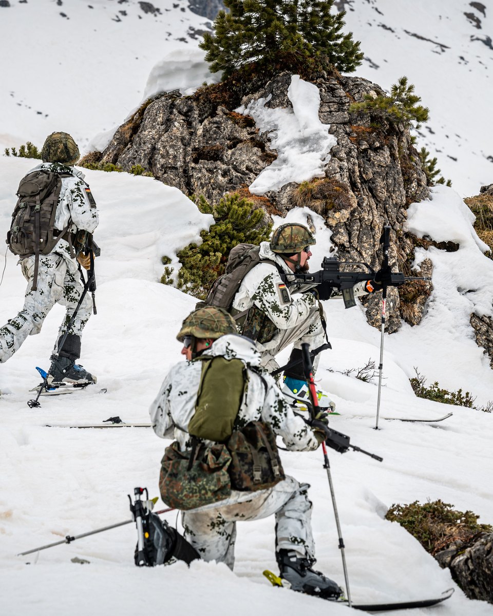 Soldaten in Schneetarn mit Gewehren gehen im verschneiten Gelände vor, einer legt vor einem Felsblock an, einer kniet im Vordergrund