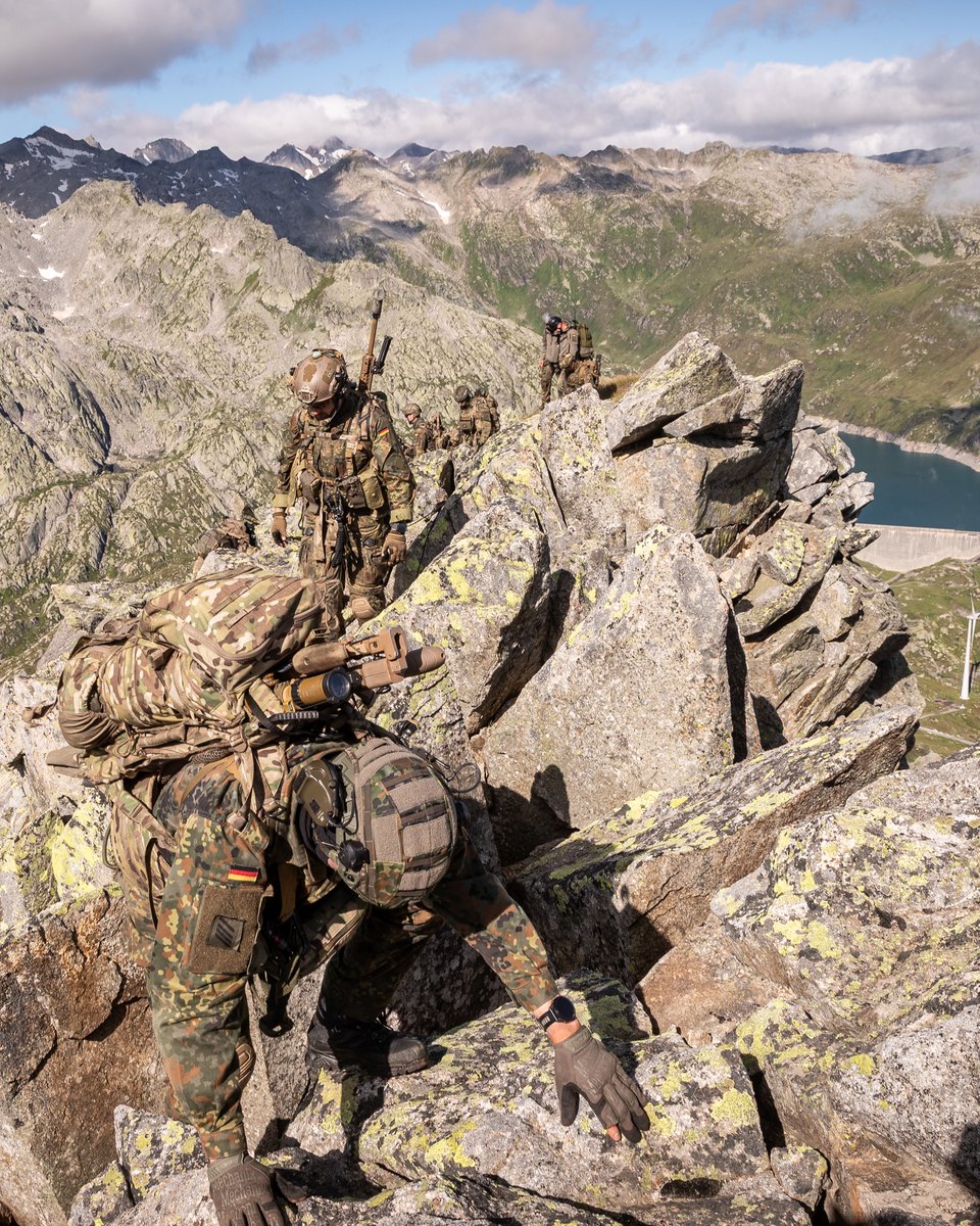 Soldaten in Flecktarn mit Helm und Gewehr auf dem Rücken steigen über einen Felsgrat, tief unten ein Stausee