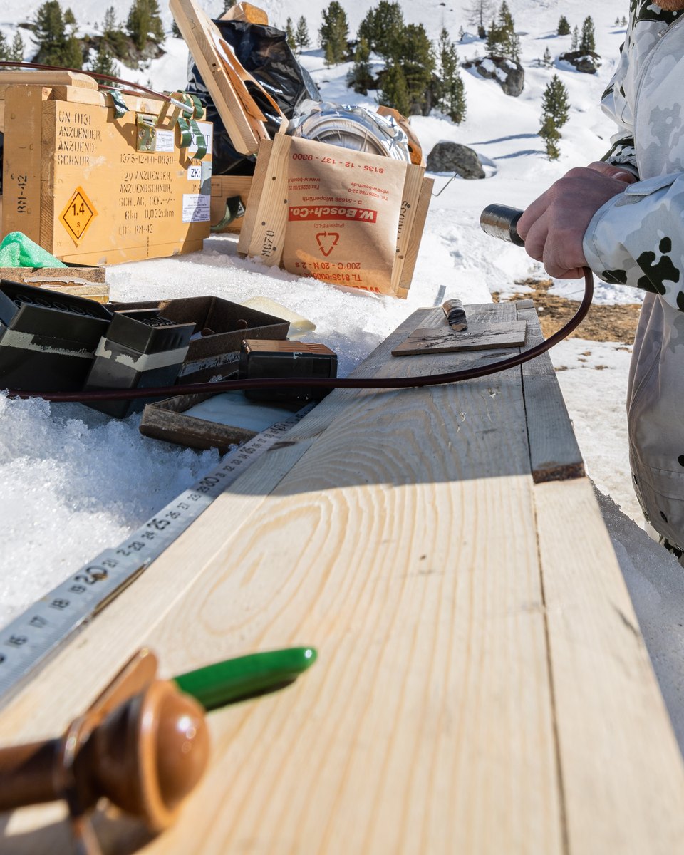 Hand an einer Anzündschnur über einem Holzbrett mit Zollstock, dahinter Kisten mit Gefahrzeichen