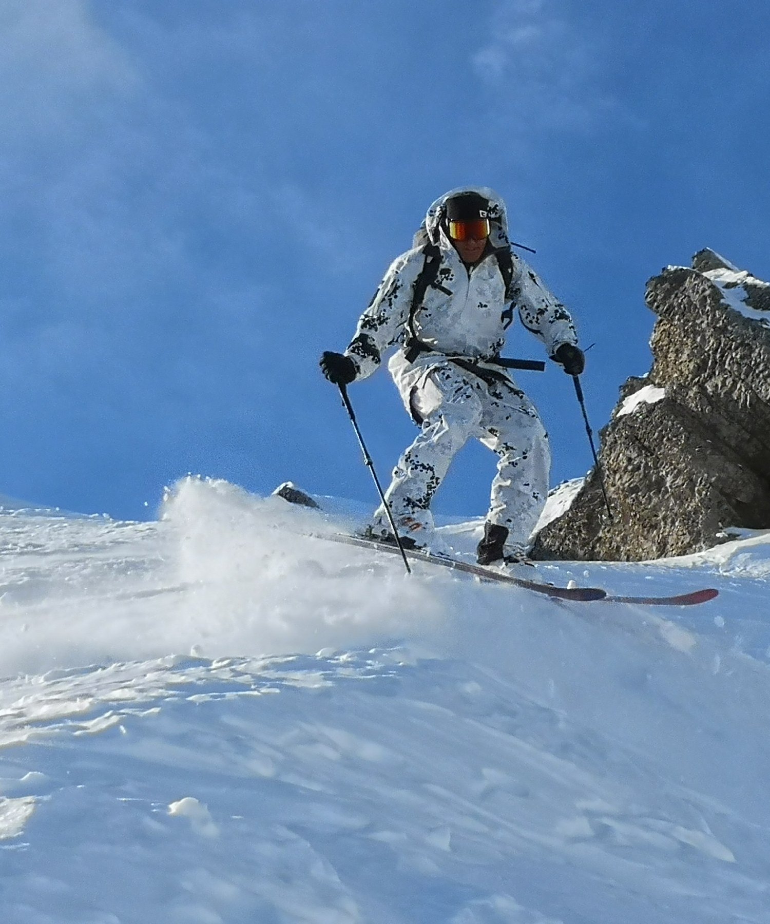 Soldat in Weißtarn springt mit Ski über eine Schneekante, Schnee stiebt auf