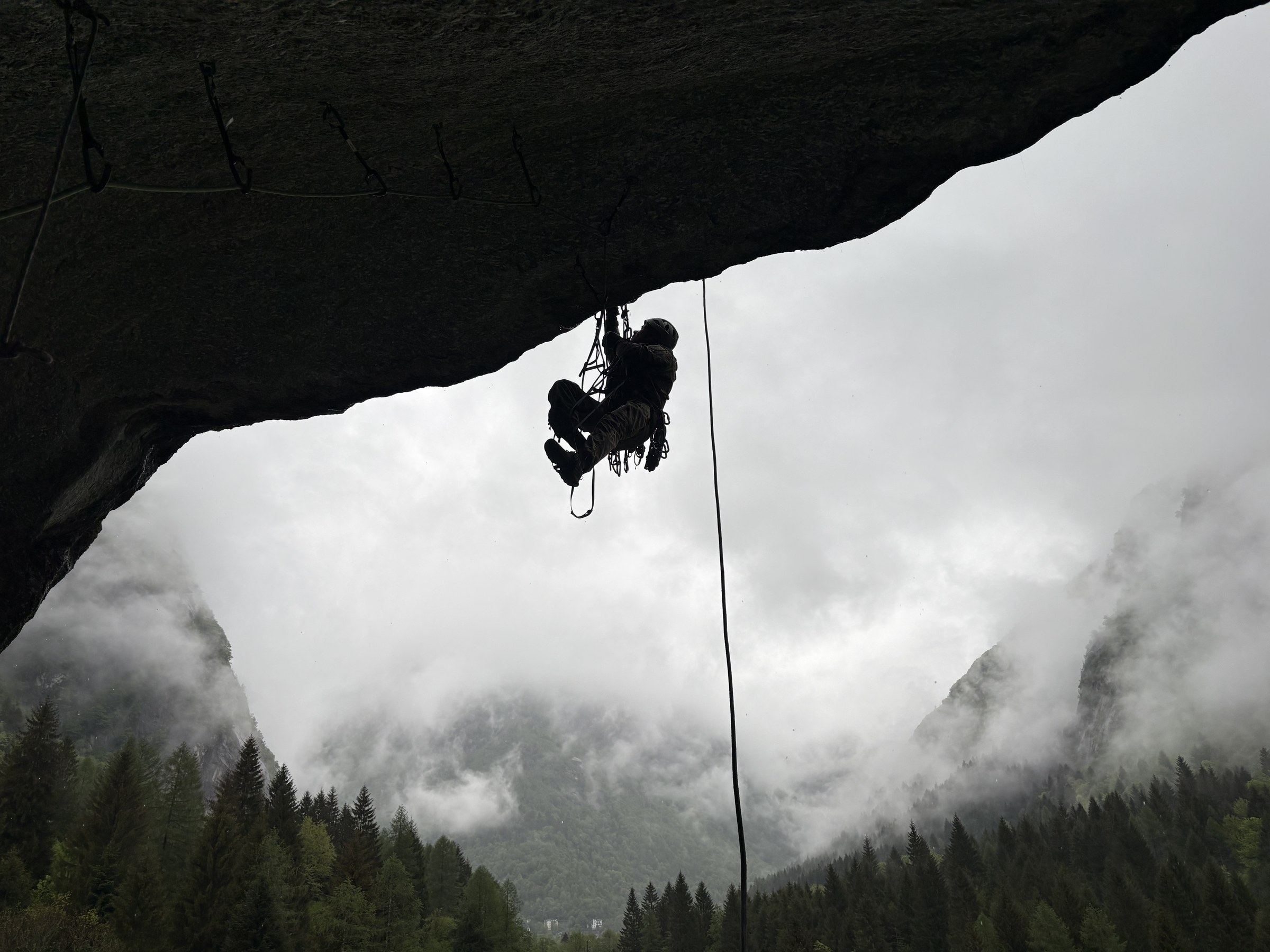 Ein Kletterer hängt in Trittleitern unter einem Felsdach, darunter Wolken im Tal