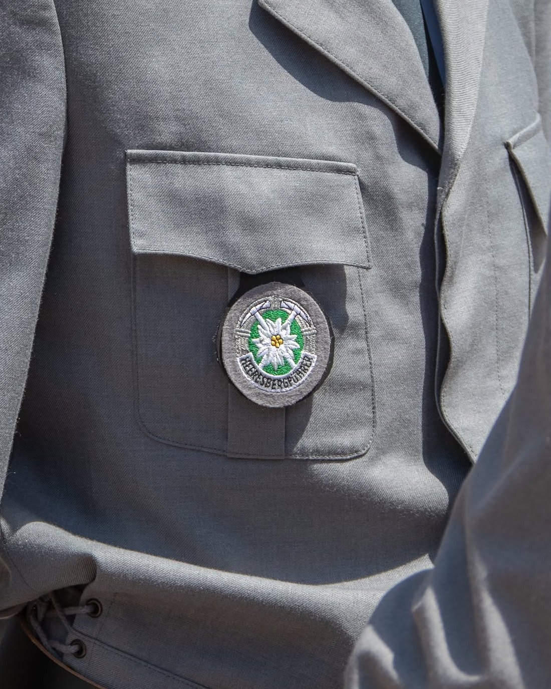 Das gestickte Heeresbergführerabzeichen mit Edelweiß, gekreuzten Eispickeln und Schriftband auf der Brusttasche einer grauen Uniformjacke
