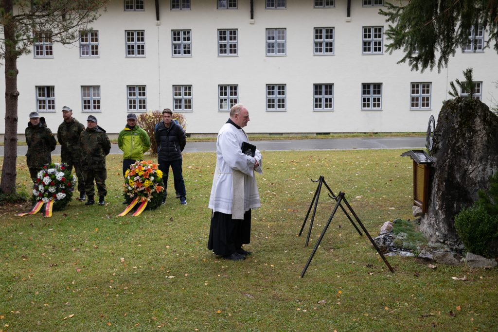 Kranzniederlegung: Geistlicher spricht am Gedenkstein, daneben zwei Kränze und Teilnehmer