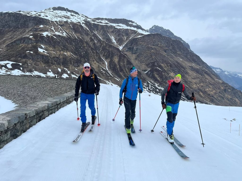 Drei Skitourengeher gehen über die schneebedeckte Staumauer, dahinter schroffe Berghänge