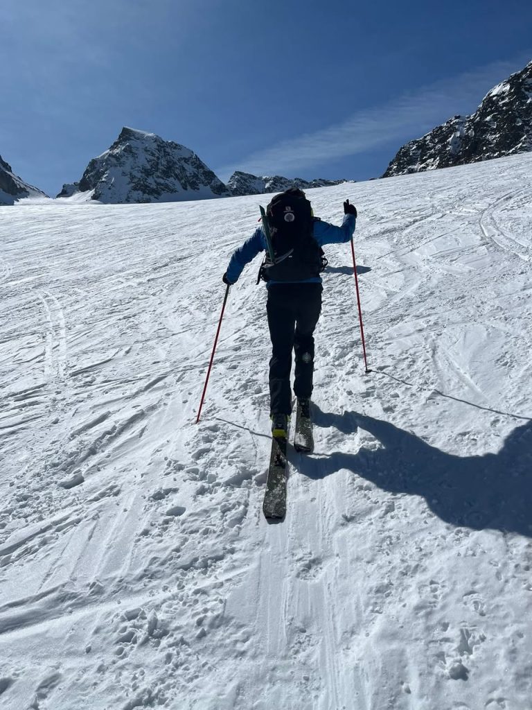 Skitourengeher steigt bei blauem Himmel einen Gletscherhang auf, im Hintergrund Gipfel