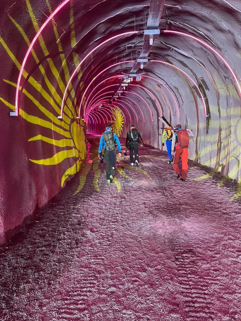 Skitourengeher tragen ihre Ski durch einen farbig beleuchteten Tunnel mit Sonnenmotiv