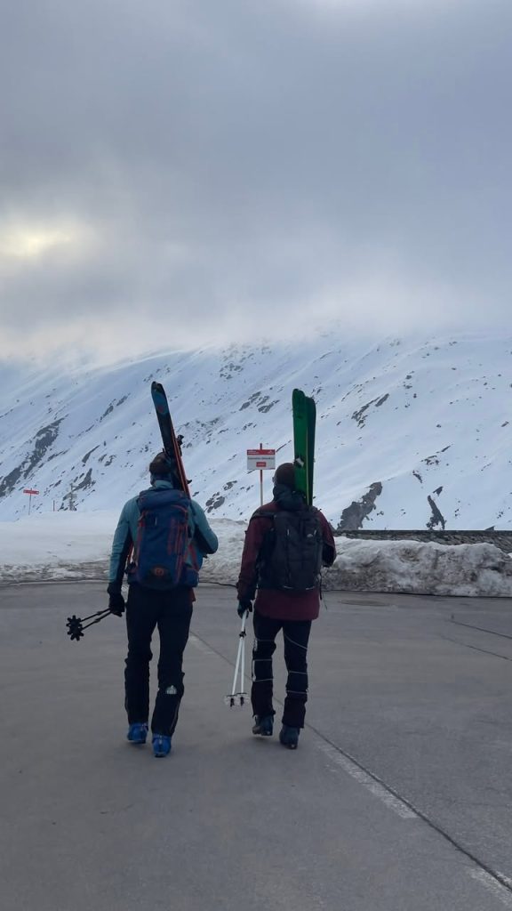 Zwei Skitourengeher tragen Ski auf dem Rücken über einen Parkplatz Richtung Berge