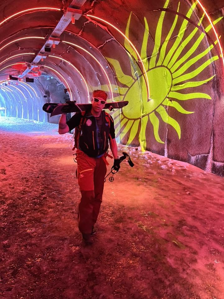 Skitourengeher mit Ski auf der Schulter geht durch einen rot beleuchteten Tunnel