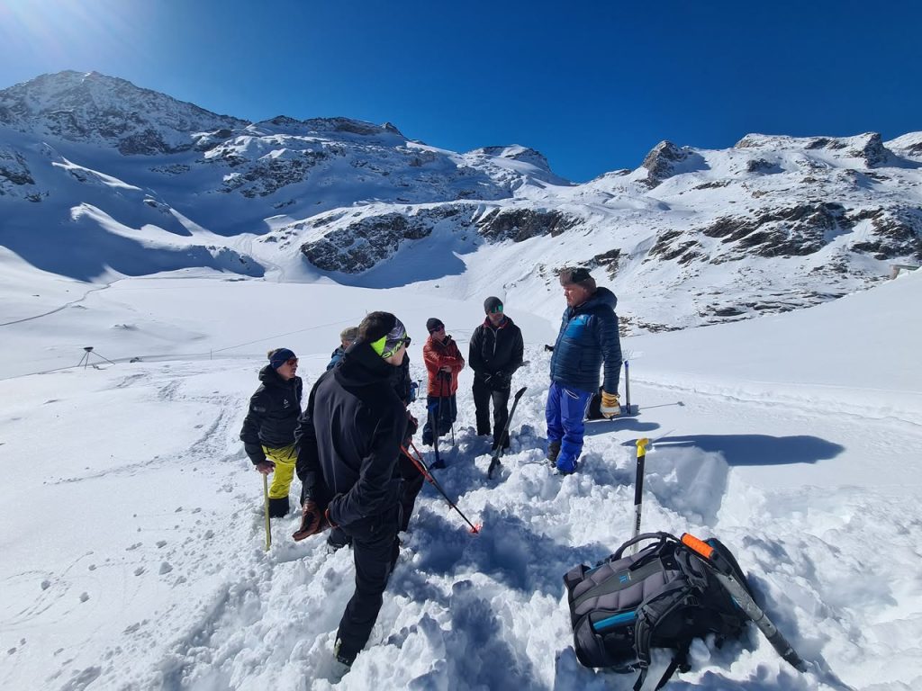 Gruppe bespricht sich am Schneeprofil im Hang, dahinter Berge im Sonnenschein