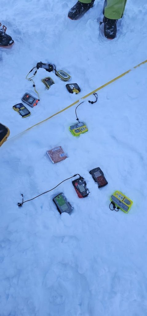 Verschiedene LVS-Geräte liegen nebeneinander im Schnee neben einem gelben Maßband